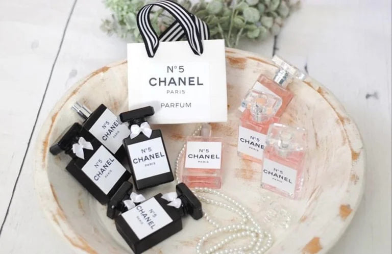 Miniature Chanel No. 5 newborn prop bottles in black and clear/pink with pearl necklace and Chanel gift bag sitting in bowl.